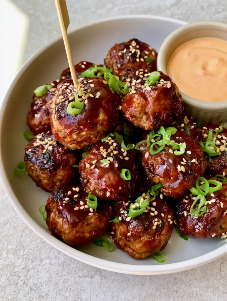 Korean BBQ Meatballs with a Thick Glossy Gochujang Glaze 2 High-angle view of Korean BBQ Meatballs in a shallow white ceramic bowl with a pale orange dipping sauce and a wooden toothpick.