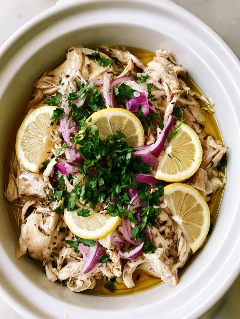 Slow Cooker Greek Chicken with Lemon and Oregano 1 Top-down view of tender Slow Cooker Greek Chicken arranged in a ceramic pot with fresh garnishes.